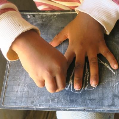 A kindergarten child traces her fingers with chalk on a blackboard to develop motor skills during a creative lesson taught by Projects Abroad Childcare volunteers in Madagascar. A child outlines her hand with chalk during a skills development lesson with a volunteer working with children in Madagascar.